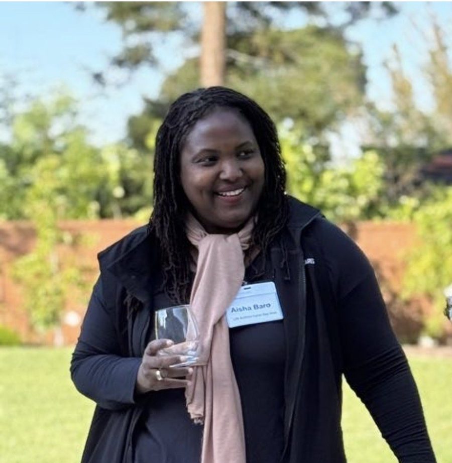 Aisha Baro smiling outdoors at an event, wearing a black jacket, a pink scarf and a name badge.