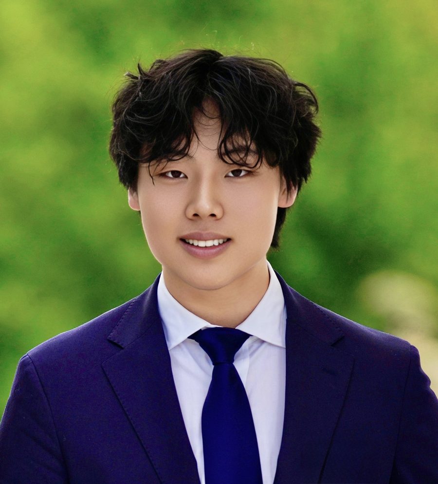 David Liu in a navy suit, white shirt and dark blue tie, photographed outdoors against blurred green foliage.