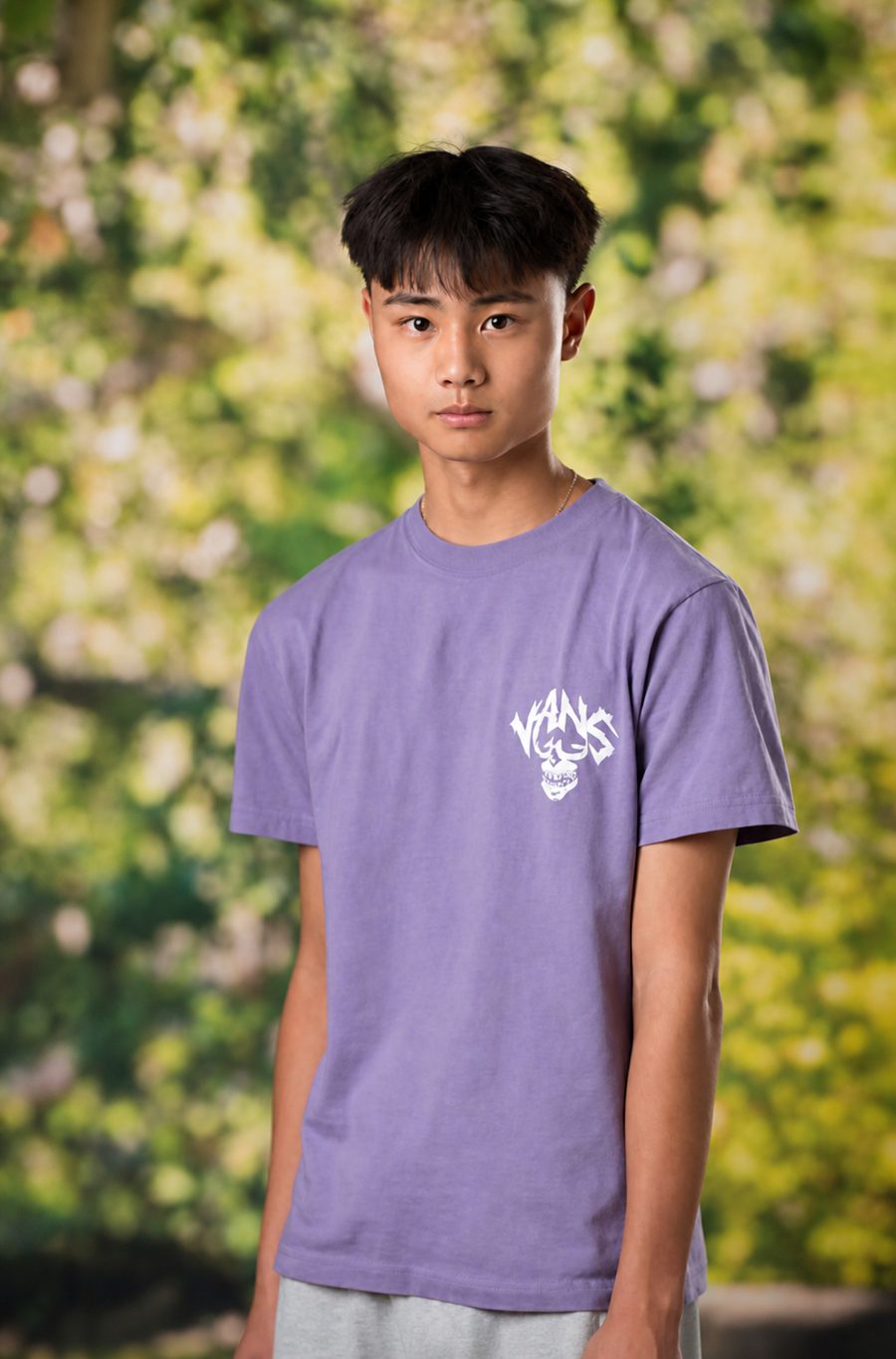 Ethan Chen standing outdoors in a lavender T-shirt in front of a wall of pale flowering greenery.