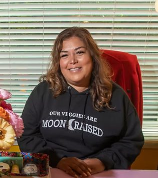 Belinda Hernandez-Arriaga seated at a desk in a dark hooded sweatshirt, with flowers and framed pictures beside her.
