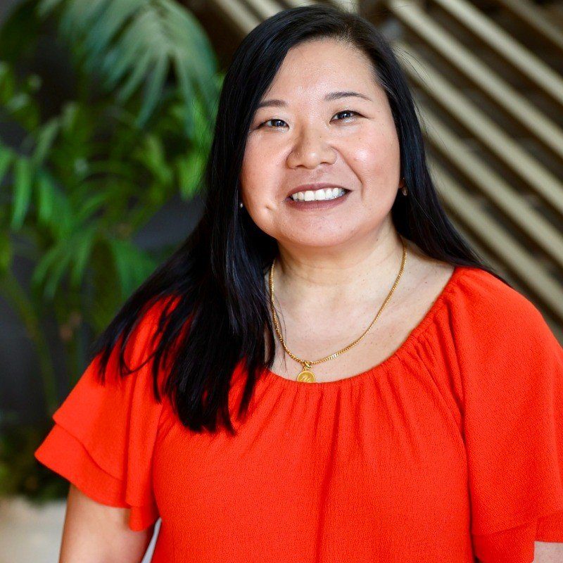 Phoebe Shin Venkat smiling in a red top, photographed indoors in front of a plant and a slatted wooden wall.