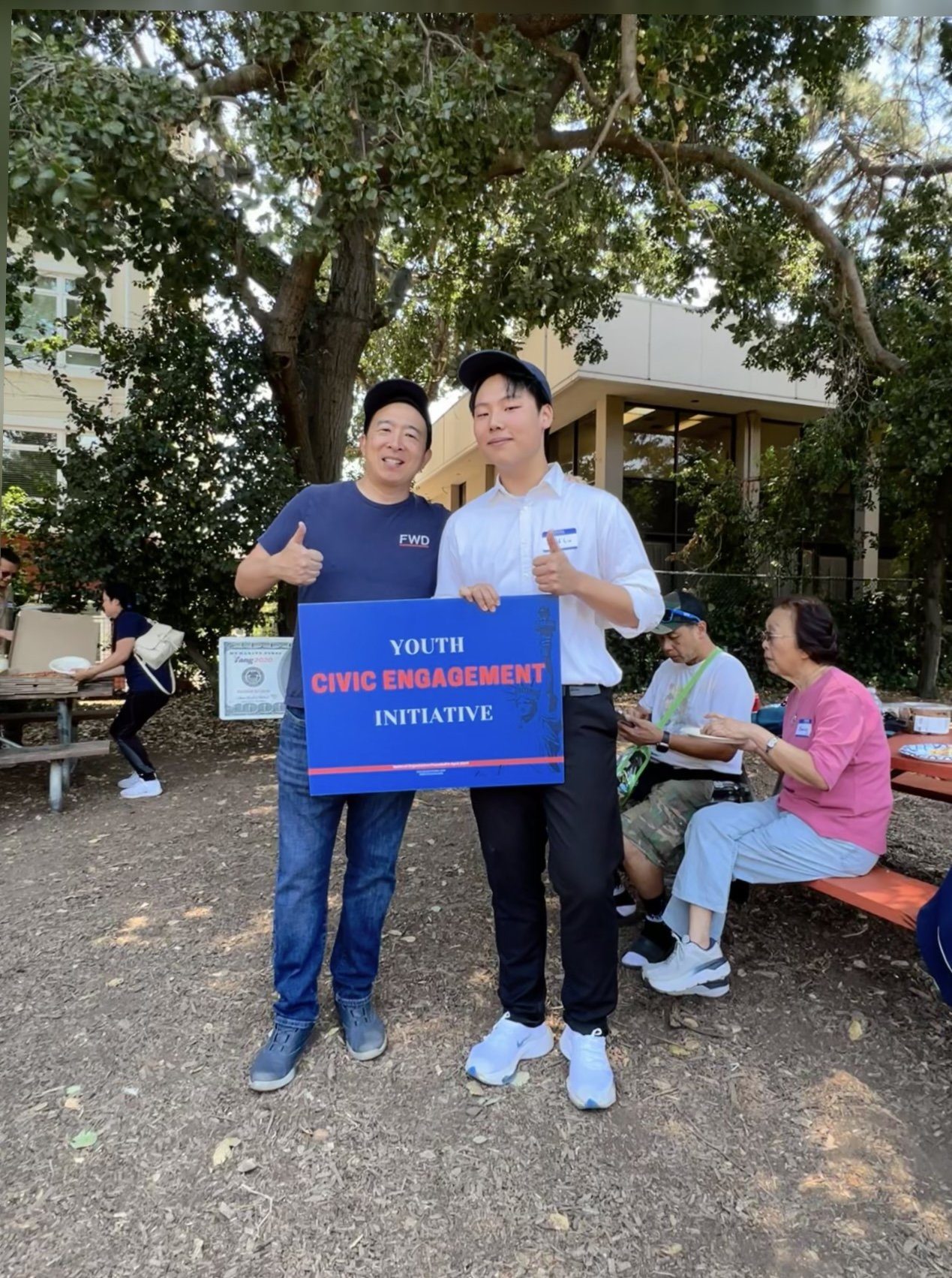 A student and Andrew Yang giving a thumbs up while holding a Youth Civic Engagement Initiative sign at an outdoor gathering with picnic tables.