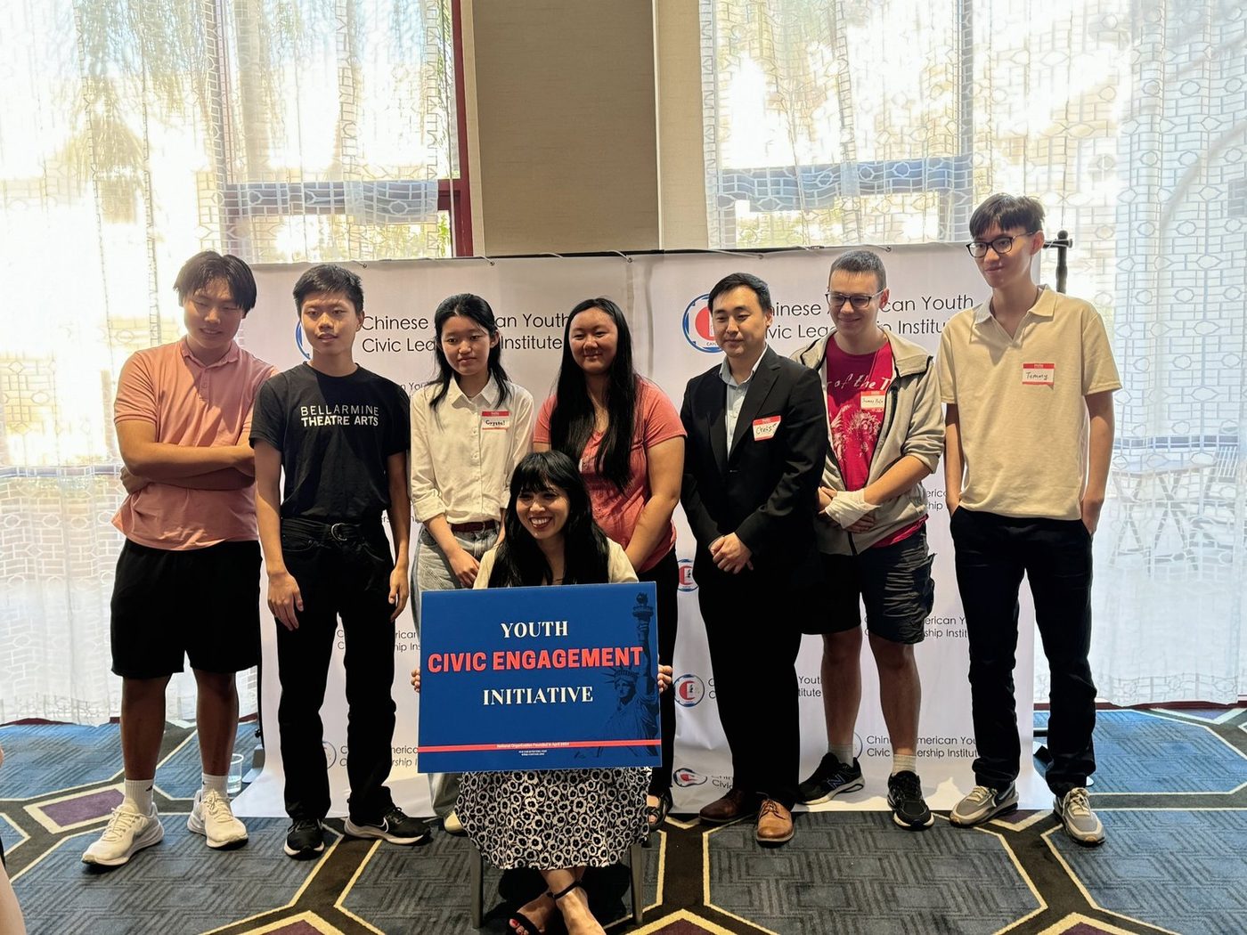 Seven people standing behind a seated young woman who holds a large blue sign reading Youth Civic Engagement Initiative, in front of a printed backdrop for the Chinese American Youth Civic Leadership Institute.