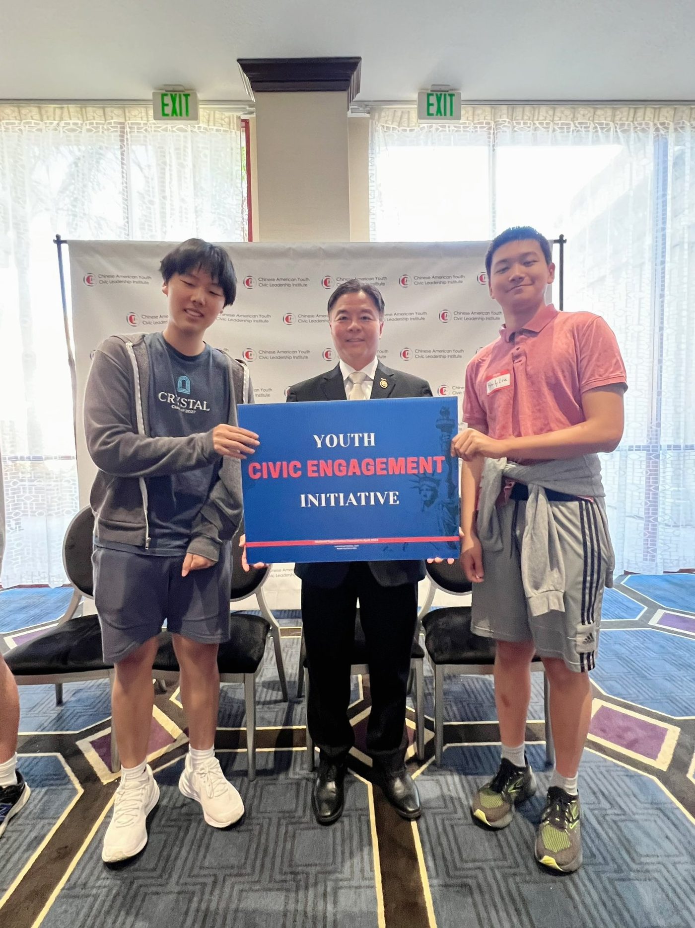 Two students holding a Youth Civic Engagement Initiative sign on either side of a man in a dark suit, in front of a Chinese American Youth Civic Leadership Institute backdrop.
