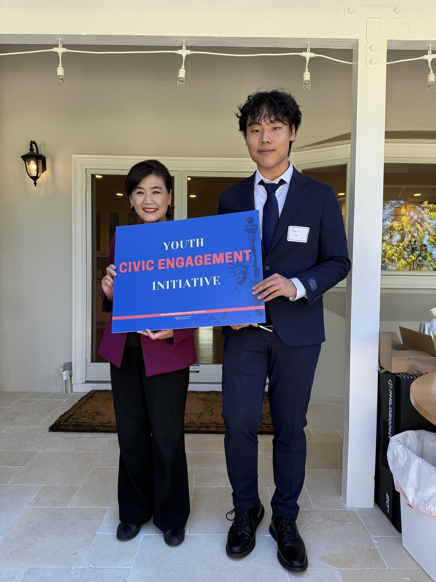 A student in a suit and Congresswoman Judy Chu holding a Youth Civic Engagement Initiative sign on a terrace.