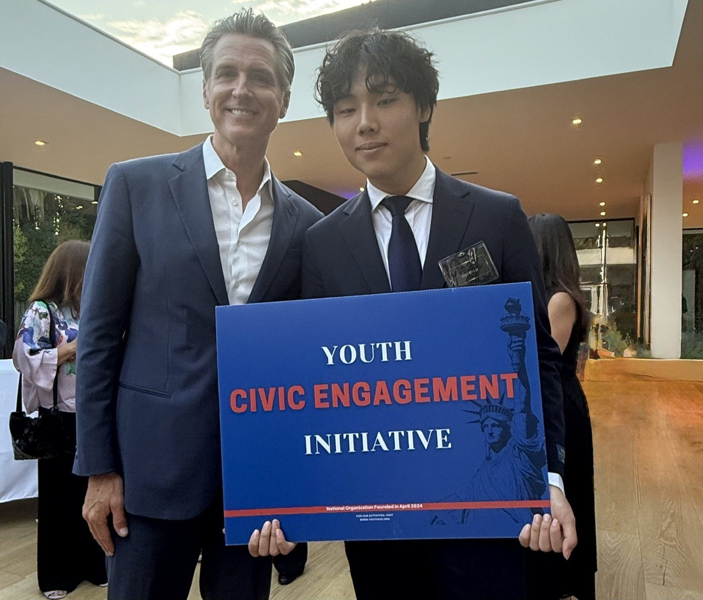 A student in a suit holding a Youth Civic Engagement Initiative sign beside Governor Gavin Newsom at an evening reception.