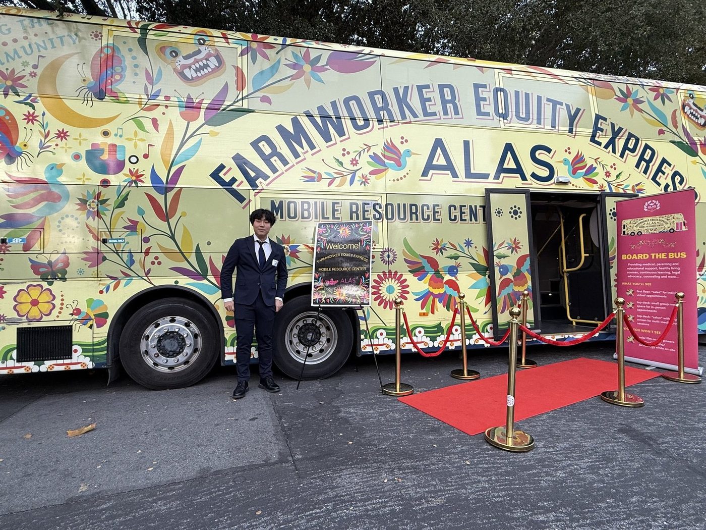 A student in a suit standing beside the Farmworker Health Equity Express, a brightly painted double decker bus lettered ALAS Mobile Resource Center, with a red carpet and rope barrier laid at its open door.