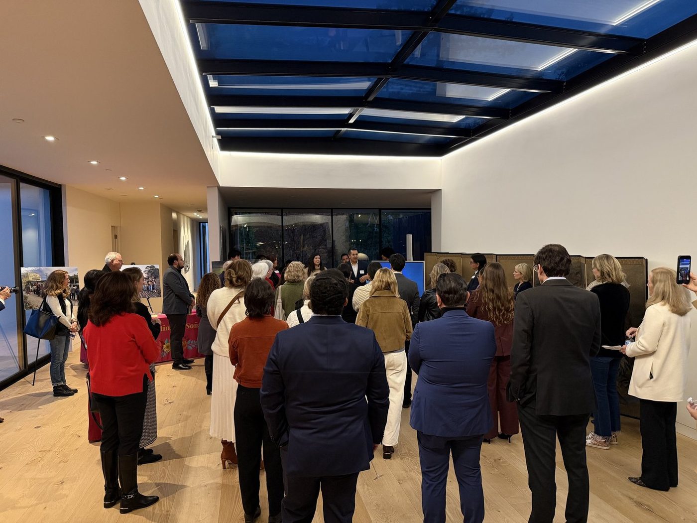 Guests standing indoors at an evening reception, listening to a speaker beside display boards of photographs of the bus.