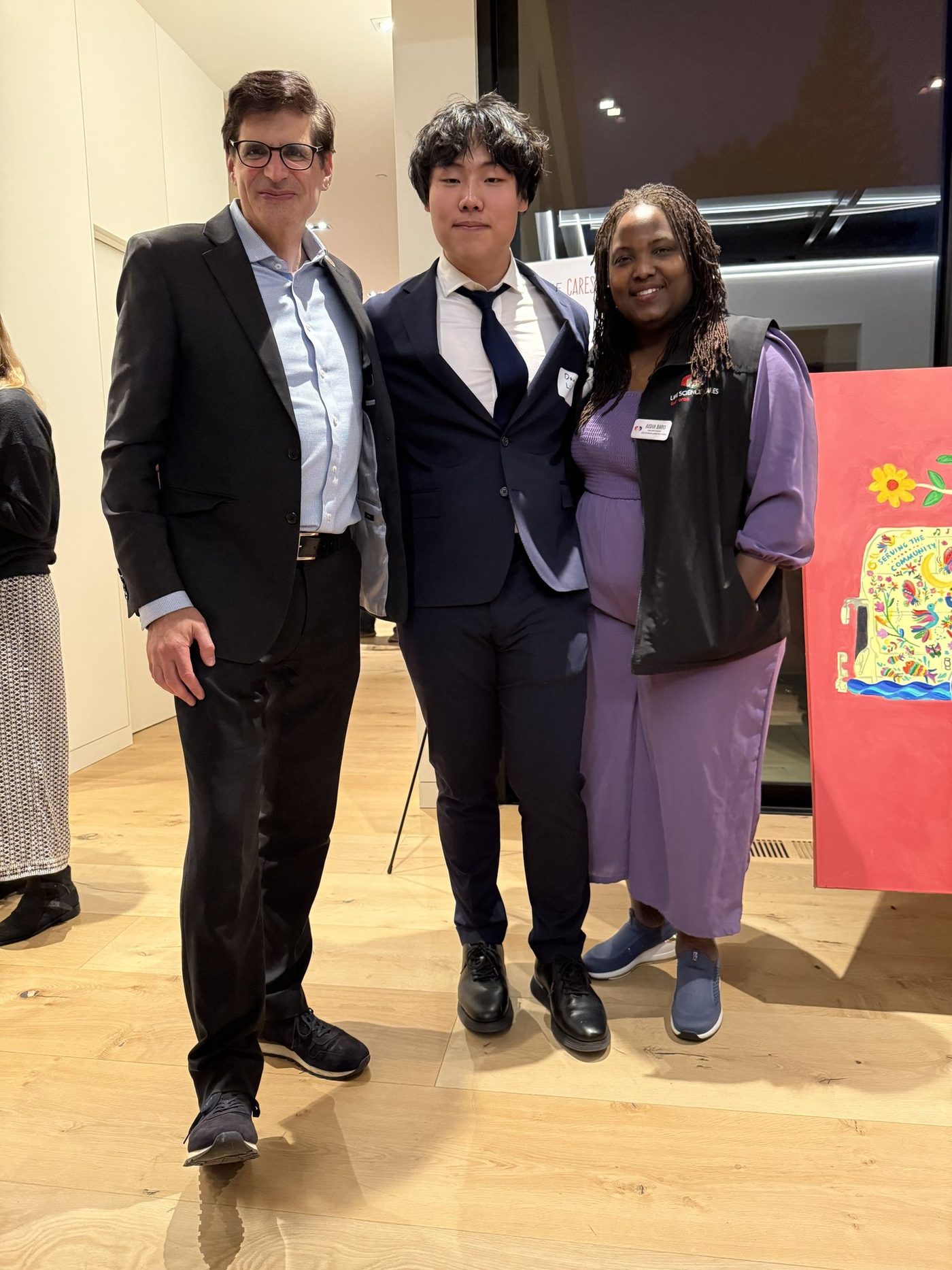 Three people standing indoors at an evening reception: a man in a dark jacket, a student in a navy suit and tie, and a woman in a purple dress wearing a Life Science Cares Bay Area vest and a name badge reading Aisha Baro, beside a painted panel of a flowered bus.