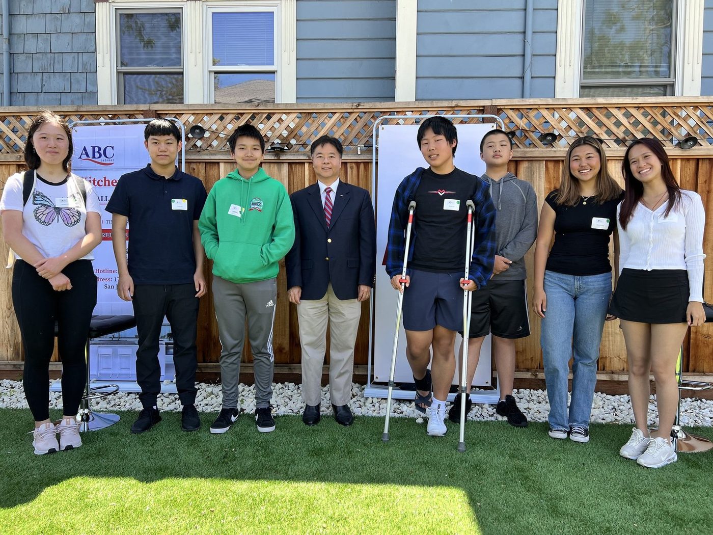 Students standing in a row with a man in a navy blazer in front of a wooden fence and a lattice screen in a garden.