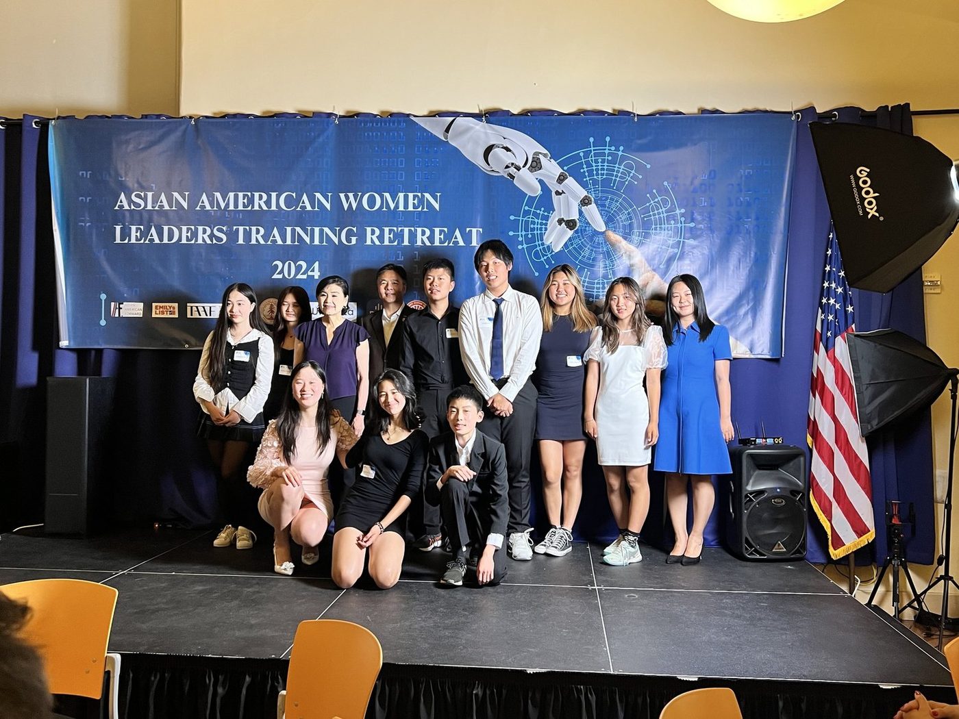 Students and adults posed on a low stage in front of a banner reading Asian American Women Leaders Training Retreat 2024, beside a United States flag.