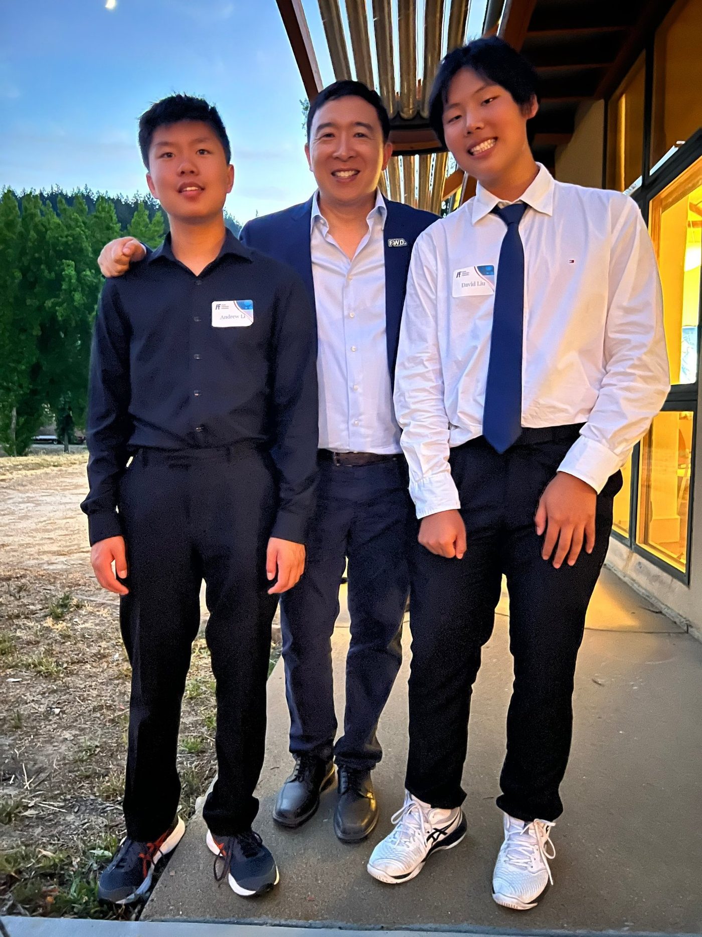 Two students in dress shirts standing on either side of Andrew Yang outside a building at dusk.