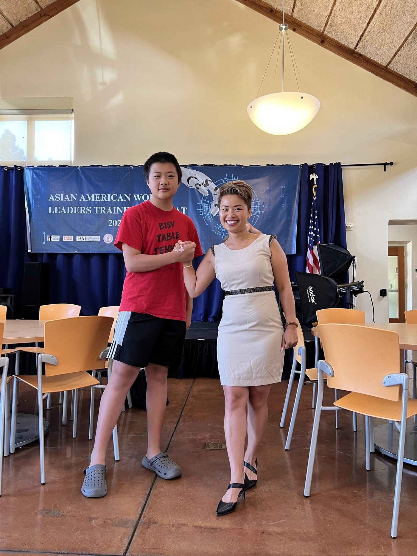 A student shaking hands with a woman in a hall, in front of the Asian American Women Leaders Training Retreat 2024 banner.
