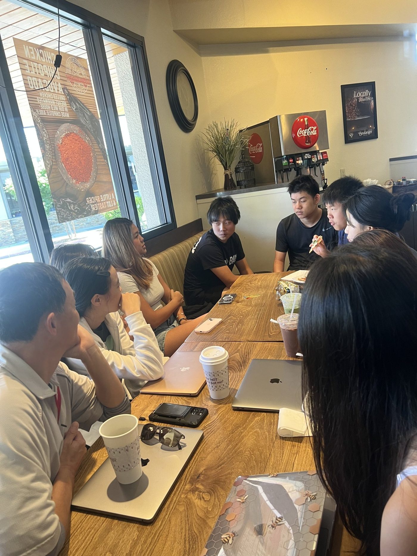 About ten students and two adults sitting around a long wooden table in a cafe, with laptops, phones and drinks in front of them.